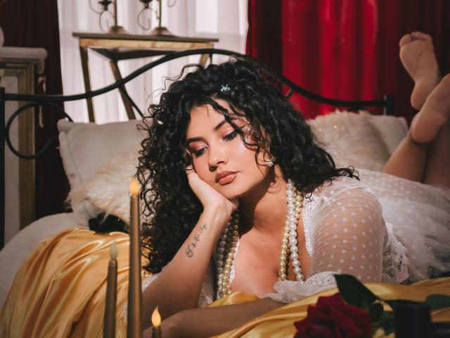 CanelaLebrad with curly black hair, lying on a bed, wearing a sheer polka-dot top, pearl necklace, and gold sheets, head on hand, looking down, tattoos visible on her wrist.