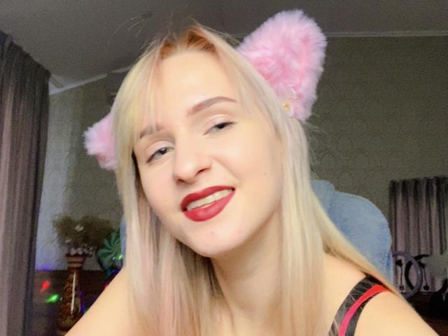 FlyFreedom with blonde hair, pink furry cat ears, red lipstick, smiling, wearing a black and red top, in a selfie, showing off her cute face and sexy look.
