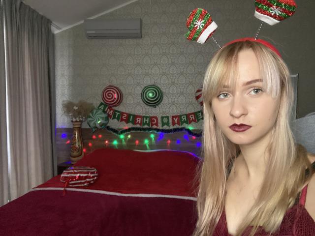 FlyFreedom has blonde hair, wearing red headband with Christmas antlers, red lipstick, and a red dress, standing in a room with Christmas decorations and colorful lights.