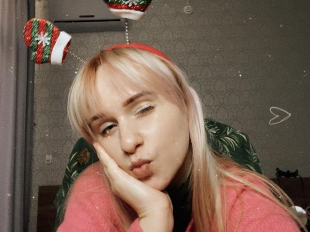 FlyFreedom with blonde hair, wearing Christmas headband with red pom-poms, pouting with hand on cheek, in pink sweater, looking cute, in selfie with subtle filter.