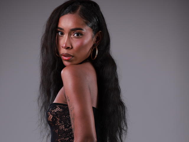 ScalettHoney with long black hair, wearing a black lace top, looking over her shoulder, showing her tats, and sporting gold hoop earrings, with a sexy, confident look.