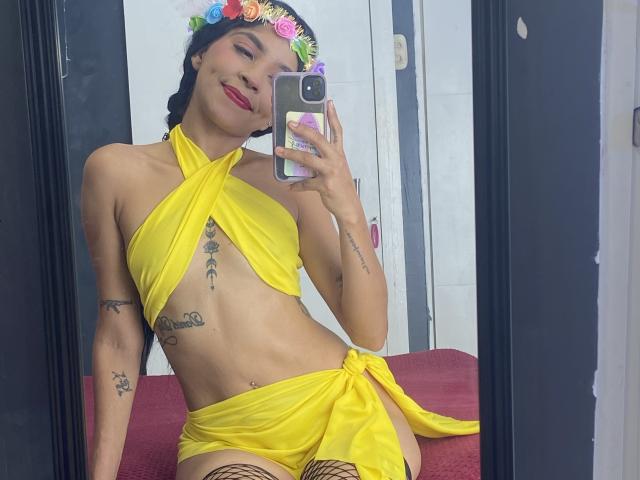 RosieRousse in a yellow wrap dress, showing her tits and pussy, with tattoos, a flower crown, red lipstick, and fishnet stockings, taking a mirror selfie.