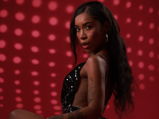 ScalettHoney with dark skin, long wavy black hair, wearing a black sequined top, looking over her shoulder, showing tattoos on her right arm, wearing large hoop earrings.