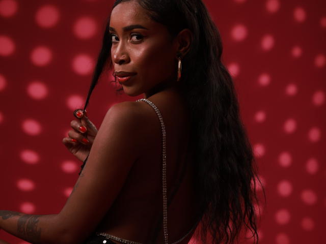 ScalettHoney with dark brown skin, long wavy black hair, red nails, holding a hair strand, looking over shoulder, wearing a silver backless dress, red earrings, subtle smile.