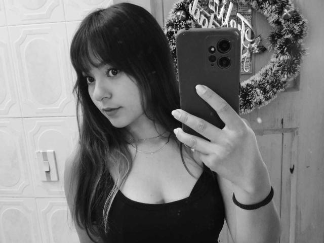 PaulinaaSweet with long dark hair, bangs, nose ring, black tank top, holding iPhone, taking mirror selfie, showing small tits, looking at camera, in black and white.