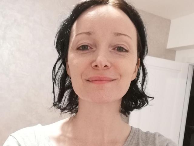 BlueFlowerr has pale skin, black wavy hair, green eyes, and a slight smile. She's wearing a grey shirt, showing a subtle neck and collarbone.