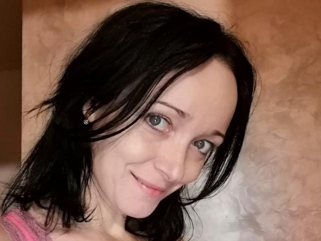 Close-up of BlueFlowerr, a woman with pale skin and black hair. She has blue eyes, a slight smile, and is wearing a pink top. Her hair is slightly messy, and she has a small earring in her left ear. The texture of the background is rough and brownish, contrasting with her smooth skin and dark hair. The composition focuses on her face, showing a casual, relaxed demeanor. Her eyes are slightly off-center, adding a dynamic feel to the image.