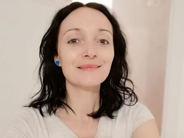 BlueFlowerr has black wavy hair, blue earrings, and a slight smile, wearing a white shirt, with fair skin and brown eyes. She's a cute, petite chick.