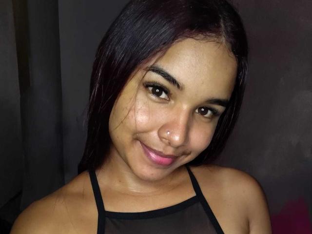 VaneIsabel tiene una piel marrón media, cabello lacio y oscuro, un pequeño aro en la nariz y está sonriendo con lápiz labial rosa. Lleva un top negro con tirantes, luciendo linda y atractiva..