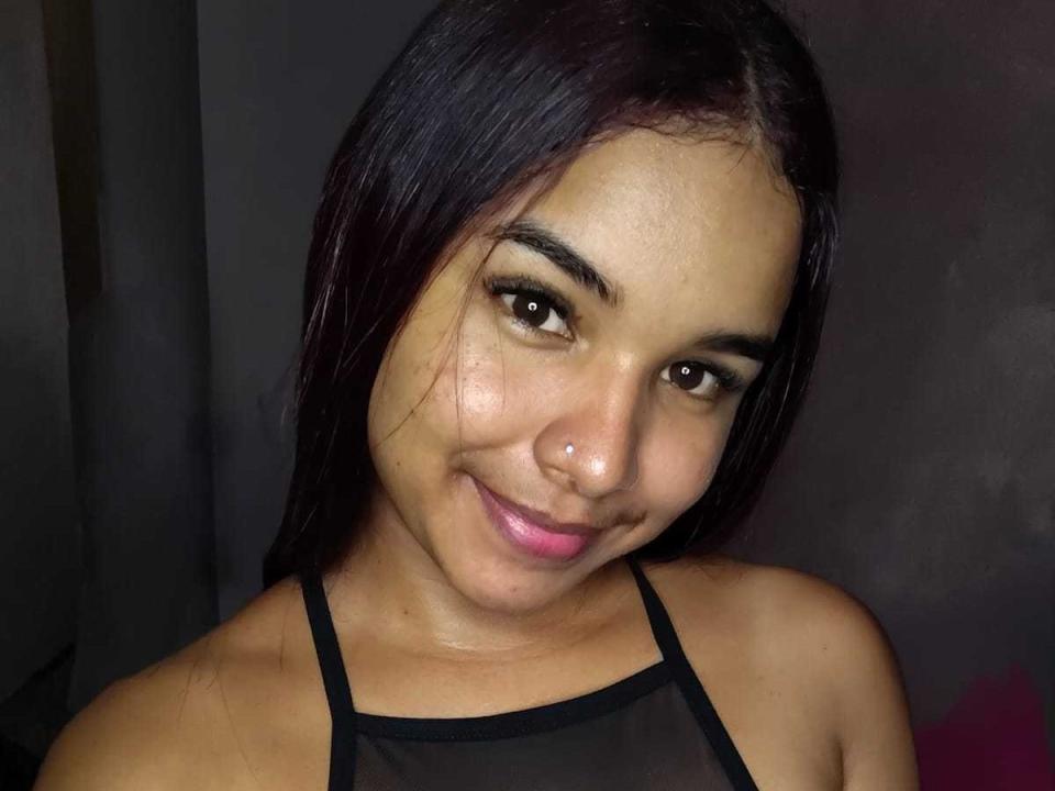 VaneIsabel tiene una piel marrón media, cabello lacio y oscuro, un pequeño aro en la nariz y está sonriendo con lápiz labial rosa. Lleva un top negro con tirantes, luciendo linda y atractiva..