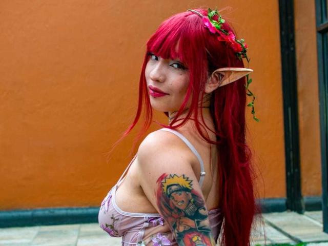 KuroBabee with red hair, elf ears, and a flower crown, looking over her shoulder, showing her tits and colorful tattoo on her right arm, wearing a pink dress.