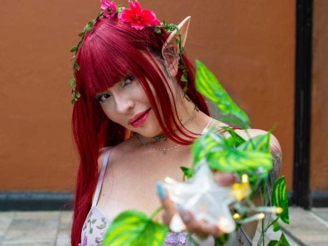 A photograph of KuroBabee with red hair, elf ears, and a floral headpiece. She's holding a green leafy plant with a white star-shaped flower. She has a subtle smile, pink lipstick, and is wearing a light-colored, lace-patterned top. Her arm has a visible tattoo. The background is a plain, brown wall. The plant is in the foreground, slightly blurred. Her hair is straight and long, with bangs. She has a fair skin tone.