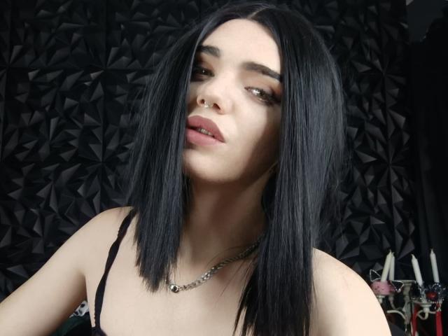 VanessaDomy with black straight hair, nose ring, and subtle makeup, wearing a black strap top, necklace, and slight pout, looking down with a relaxed expression.