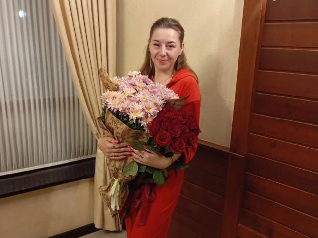 JennaMollberty with a big bouquet of pink and red flowers, holding them with both hands, wearing a red dress, smiling softly, standing next to wooden wall panels.