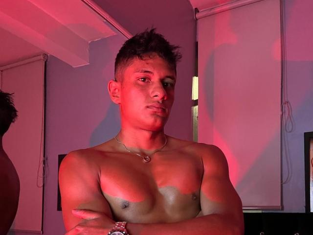 JamessHott senza camicia, con petto muscoloso, capelli ricci marroni corti, illuminazione rossa, braccia incrociate, collana e braccialetto, sguardo serio, in piedi in una stanza poco illuminata..