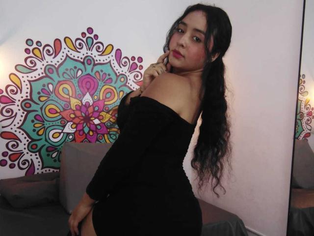 ScarlettZ with long black hair in a ponytail, wearing an off-shoulder black dress, looking over her shoulder, touching her cheek, showing her ass, in a dimly lit room.