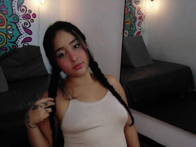 ScarlettZ with black braids, pink blush, and white tank top, showing tattoos on her right hand, sitting in front of a mirror, looking at the camera.