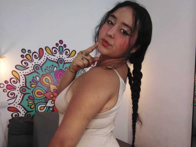 ScarlettZ with dark braids, wearing a white tank top, pointing to her pouty red lips, showing off her tits, in a slightly turned pose, with a cute, tattooed finger.