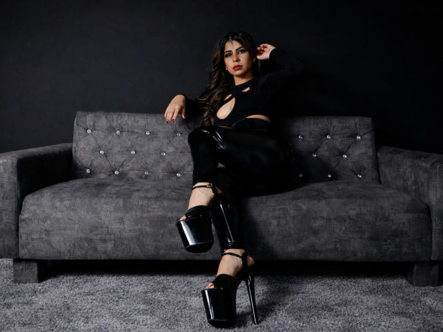 NicolBennett with long black hair, wearing a black cut-out top and shiny black pants, sitting on a gray sofa, in high-platform black heels, showing her sexy legs and butt.