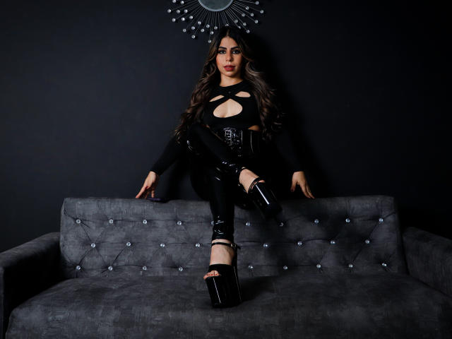 NicolBennett with long brown hair, wearing black cut-out top, shiny black pants, and platform heels, sitting on a gray tufted sofa, legs crossed, hands on arms.