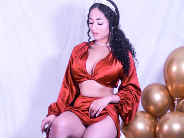 Aurora Ferretti, con cabello negro rizado, vestida con un brillante conjunto de satén rojo, mostrando sus senos, y una diadema blanca, sentada con globos dorados a su derecha..