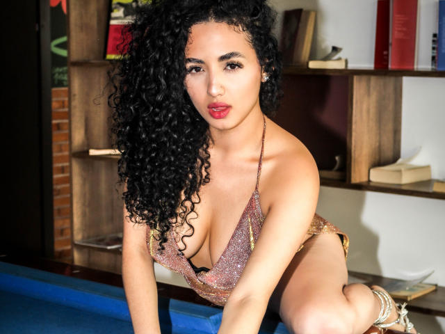 Aurora Ferretti, com cabelos pretos encaracolados, vestindo um biquíni marrom brilhante, apoiada em uma mesa de bilhar azul, exibindo seus seios grandes, brilhantes e cintilantes, e uma expressão confiante e sexy..