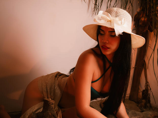 KristtieEva with dark hair, green bikini top, and tan hat with white bow, kneeling on sand, wearing a beige skirt, showing her ass and looking down with red lips.