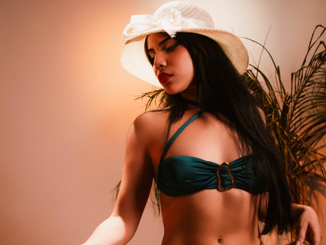 KristtieEva with long black hair, wearing a green strapless bikini top with a buckle, and a white sunhat with a bow, has her eyes closed and a pouty expression.