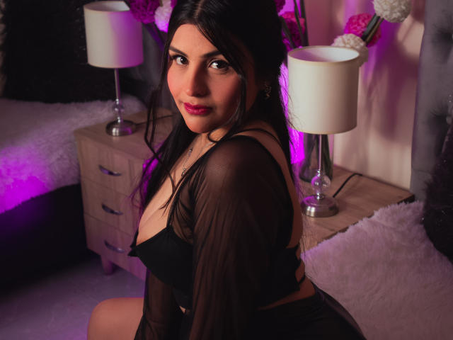 SaraWestt, con cabello oscuro, lápiz labial rosa, una blusa negra transparente que deja ver sus senos, sentada en una cama, con lámparas moradas y blancas, una mesita de noche de madera, almohadas blancas y suaves, y una mirada seductora..