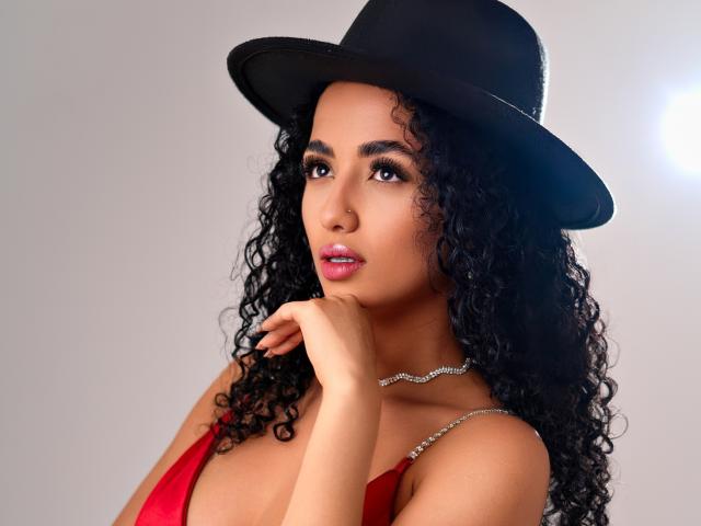 Aurora Ferretti, con cabello negro rizado, usando un sombrero negro, un vestido rojo y un collar de plata, con su mano cerca de sus labios rosados, con sus senos visibles, sobre un fondo gris..