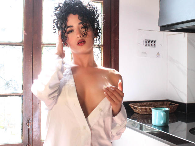 AuroraFerretti with curly black hair, open white shirt revealing her bare tits, sunlight through wooden window, hand in hair, other hand on chest, blue mug on black counter.