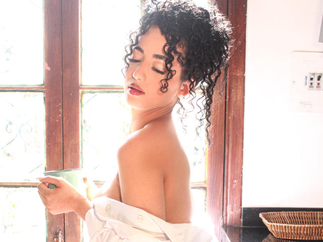 AuroraFerretti with curly black hair, red lipstick, and a white shirt slipping off her shoulders, holds a green glass, eyes closed, sunlight through wooden window.