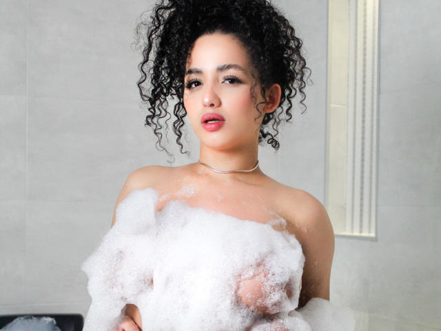 AuroraFerretti, with curly black hair, fair skin, and red lipstick, is in a bathtub covered in white bubbles, revealing her small, round tits. She wears a silver necklace.