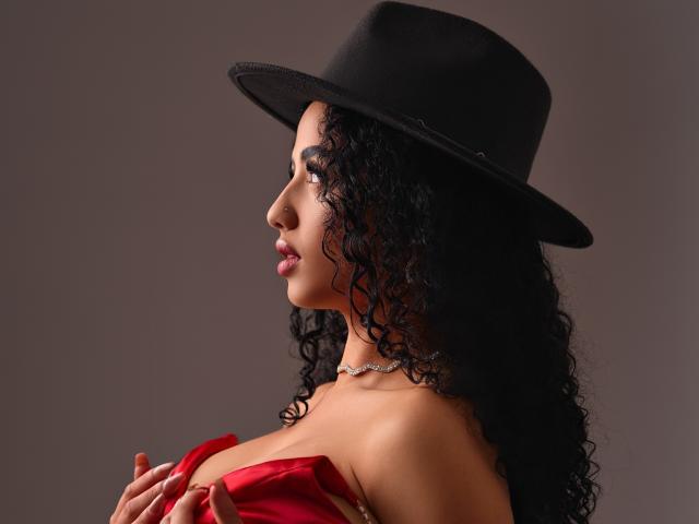 Aurora Ferretti, com cabelos pretos encaracolados, usando um chapéu preto e um vestido vermelho, segurando os seios, batom rosa, colar de choker, perfil lateral, fundo marrom escuro, sexy..