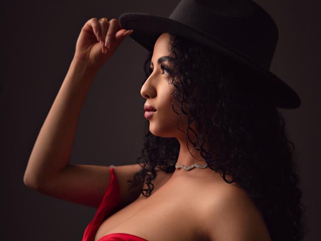 AuroraFerretti with curly black hair, wearing a black hat, red dress, and necklace, showing off her tits, holding the hat, dark background, side profile, confident look.
