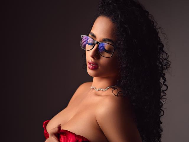 AuroraFerretti with curly black hair, wearing glasses, red strapless top, exposing her tits, red lipstick, and a silver necklace, looking to the side with a seductive expression.