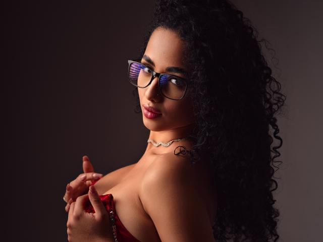 AuroraFerretti with curly black hair, wearing glasses, and a red strapless top, holding her tits with a confident smile, a necklace with a small charm. Dark background.
