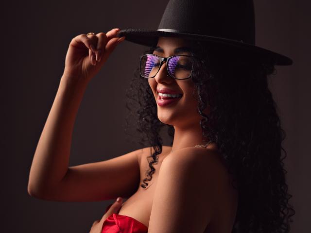 AuroraFerretti with curly black hair, wearing a black hat and purple reflection glasses, smiling, holding her hat, red strapless top, dark background, showing her tits and nose.