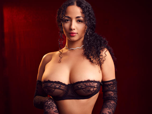 AuroraFerretti with curly black hair, wearing black lace bra and matching arm sleeves, exposing large boobs, and a silver choker, against a dark red background.