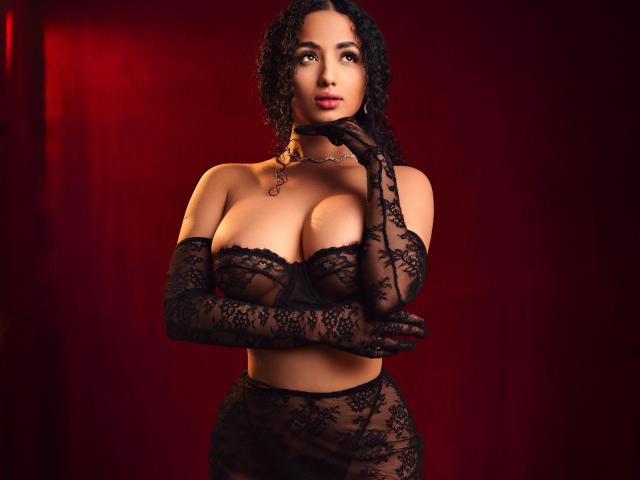 AuroraFerretti with curly black hair, wearing black lace lingerie, gloves, and choker, has large tits and a sexy, confident look, standing against a red background.