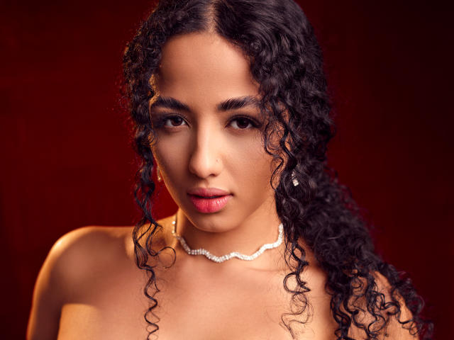 AuroraFerretti has curly black hair, brown eyes, and wears a diamond necklace against a red background. Her tits are visible, with a bare shoulder and red lips.