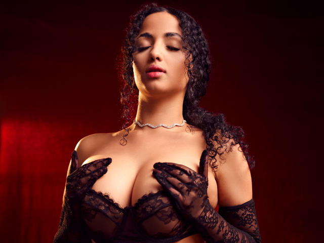 AuroraFerretti with curly black hair, wearing black lace gloves and a matching bra, covering her big, bare tits with her hands, wearing a silver necklace, red-lit background.