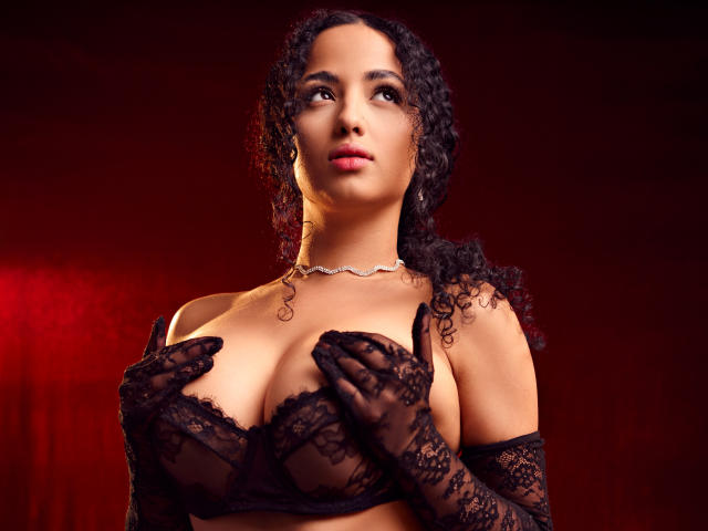 AuroraFerretti with curly black hair, wearing black lace bra and gloves, covering her big tits with her hands, red background, looking up, choker necklace, serious expression.