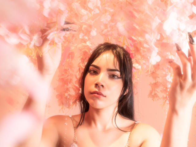 KristtieEva with dark hair, pink flowers in her hands, looking at the camera with soft makeup and a pink background. She's wearing a white top with thin straps.