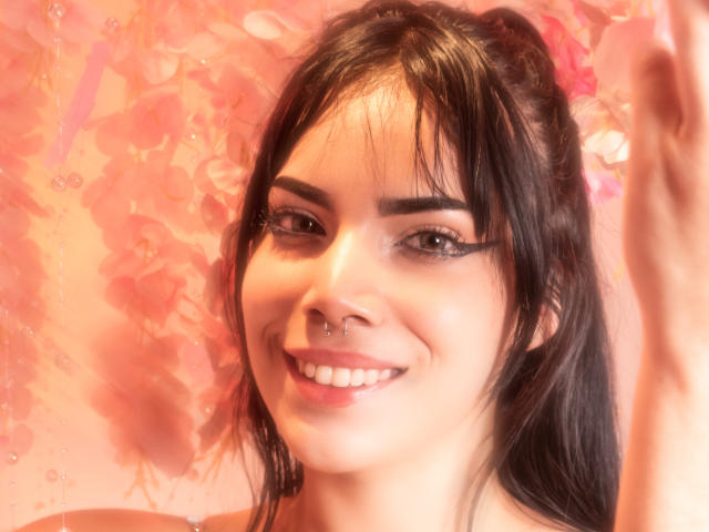 KristtieEva with dark brown hair, septum piercing, and smiling, has thick black eyeliner, and is raising her right hand. Her pinkish skin is softly lit.