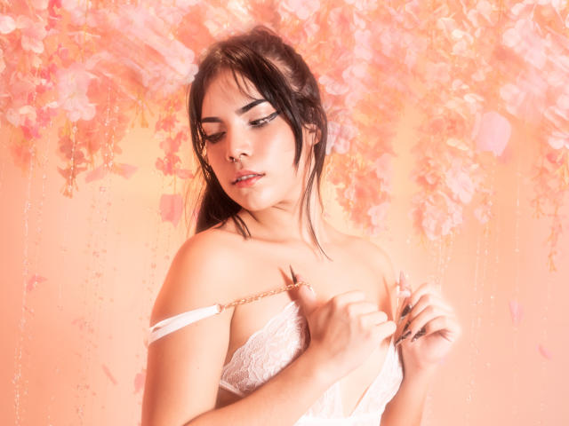 KristtieEva with dark hair, looking down, pulling down white lace bra straps, showing her tits, in a soft pink, floral, bokeh background, with a sensual, seductive expression.