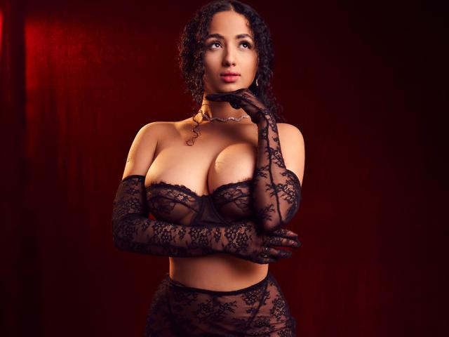 AuroraFerretti with curly black hair, wearing black lace bra, matching gloves, and choker, showing off her big tits and slim waist, against a red background.