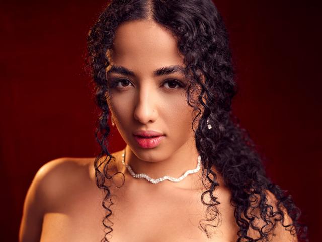AuroraFerretti has curly black hair, brown eyes, and is wearing a sparkly necklace. She's topless with medium, round tits and a serious expression. Red background.