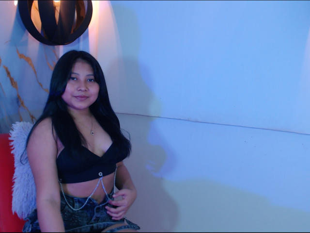 TammyGreyy with long black hair, smiling, wearing a black bralette, showing her tits, and a denim skirt with chains. She's sitting against a red and white wall.