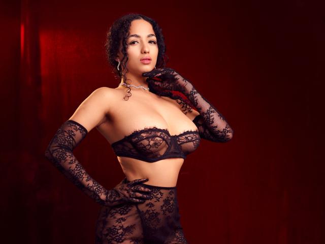 AuroraFerretti with curly black hair, wearing black lace lingerie, gloves, and a necklace, has big tits and a confident pose against a red background.